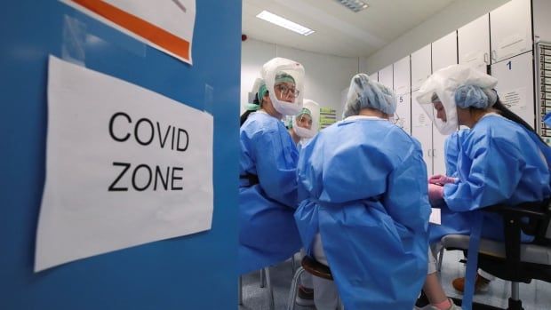 Doctors in full protective gear work in a room just past a door that says, "COVID Zone."