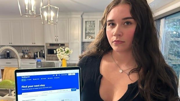 A young white woman wearing a black shirts sits at a kitchen table beside an open laptop. The words on the screen say find your next stay.