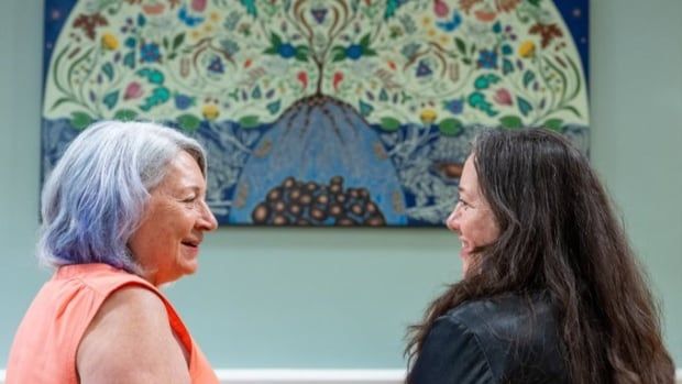 Two women stand in front of a painting looking at each other with their backs to the camera.