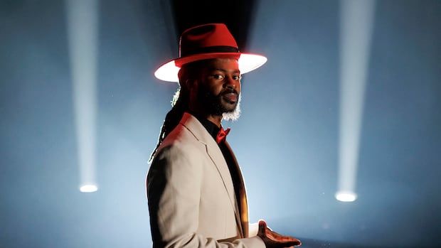 Randell Adjei has a red hat and white suit, and stares at the camera while spotlights illuminate the sky behind him.