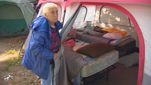 A woman with short white hair and wearing a long blue coat shows inside her tent, which holds 2 separate beds and piles of belongings. 