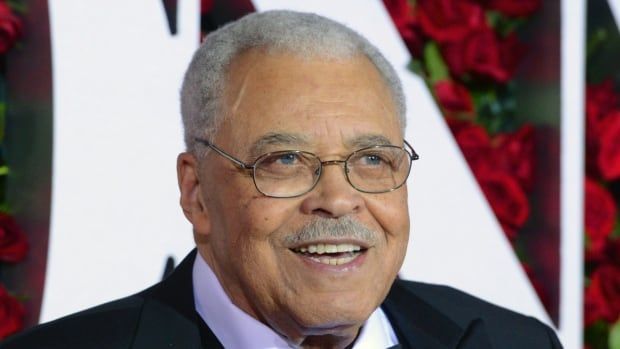 A man with grey hair, round sunglasses and a black tuxedo smiles on a red carpet.