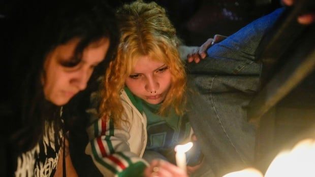 Two young women huddle closely and look mournful as one holds a candle.