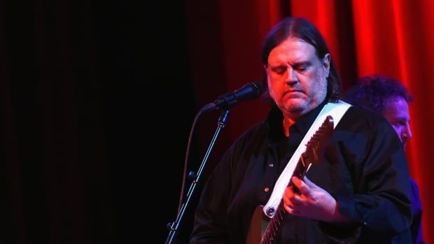 A bearded man in dark clothing plays a guitar onstage.