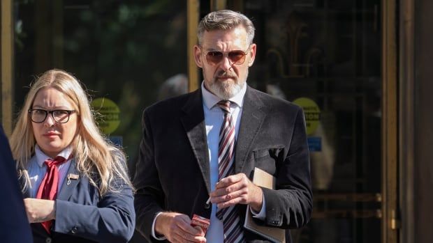 A bearded man wearing sunglasses and a suit and tie holds a cigarette while walking outside a building beside a woman. 