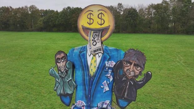 A giant effigy of a figure in a suit, with a money-face emoji, puppetting two figures that look like Oasis band members.