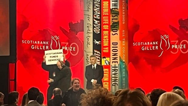 A person holds up a white sign that says "SCOTIABANK FUNDS GENOCIDE" in black letters in front of their face. Another person wearing a headset is attempted to usher them off stage. The tops of heads of a crowd is visible at the bottom of the image. At the back is a red backdrop that says Scotiabank Giller Prize. 