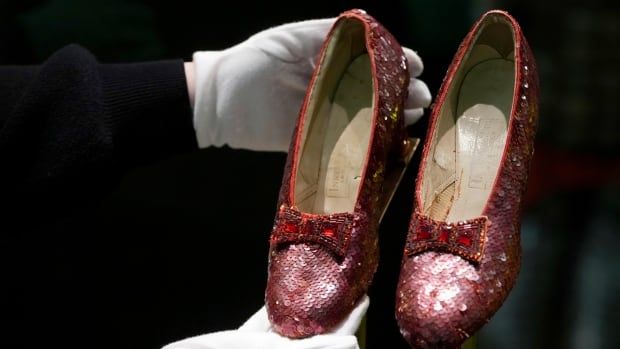 A person wearing white gloves holds up a pair of ruby slippers.