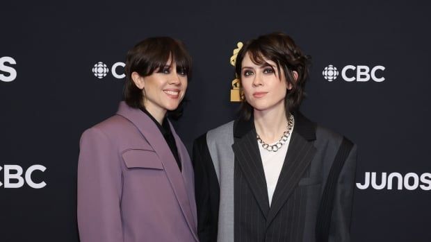 Tegan and Sara wear suits on the red carpet at the Juno Awards, posing with a black background behind them.