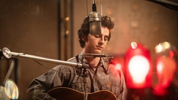 A man plays a guitar while standing in front of a microphone in a studio. 