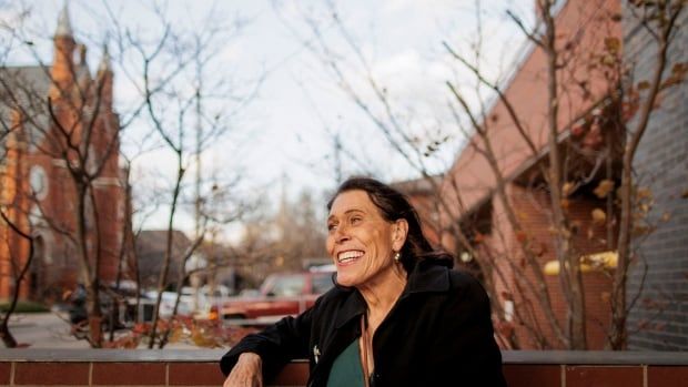 A person with long hair and a big smile stands outside in Hamilton. 