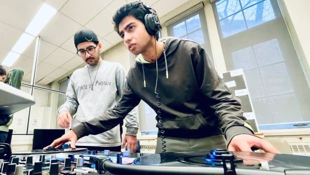 A male student wearing headphones leans over a mixing board and turntables as he looks into a laptop off-screen, while another male student to the left looks on.