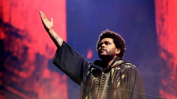 The weeknd wears a gold and black cape while raising his arm as he performs live in Las Vegas.