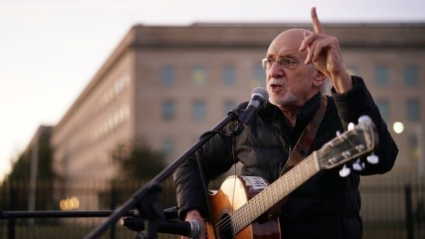 An older man standing in front of a microphone with a guitar slung around his chest is shown. The man is mid-speech, holding up one finger. 