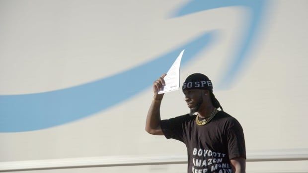 A man holds a piece of paper up to shield the sun from his eyes. He is standing in front of a massive white backdrop with a blurry blue swoop on it, and he's seen from the waist up only. 