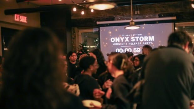 A crowd of people is photographed from within the crowd, in a room at nighttime, with muted, warm lighting. Most people are wearing black, and a screen in the distance says "Onyx Storm Midnight Release Party". 