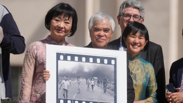 A group of people smile while holding up a photograph. In the black and white photograph they're holding, children can be seen running and crying from a smoke cloud behind them.