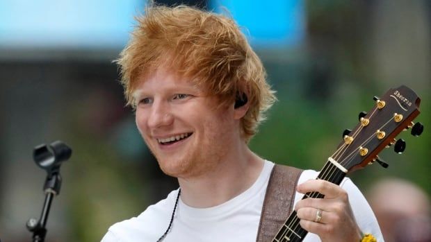 A red-haired man is shown from the shoulders up, smiling as he plays a guitar. A microphone is in front of him. 