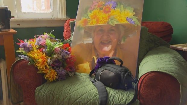 The photo of an older lady sitting on a couch. She wears a bright yellow shirt and a hat full of flowers. In the couch in front of her photo is that same hat and a camera bag. 