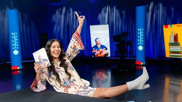 A woman with long hair leans back on her chair with a leg up and peace sign. She's holding a book and smiling.