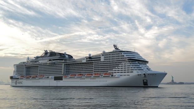 A cruise ship on the water beneath white clouds in the blue sky, with the Statue of Liberty in the distant background. 