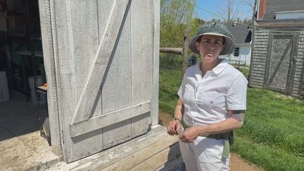 A woman, wearing a white jumpsuit and a hat, stands in front of a barn. 