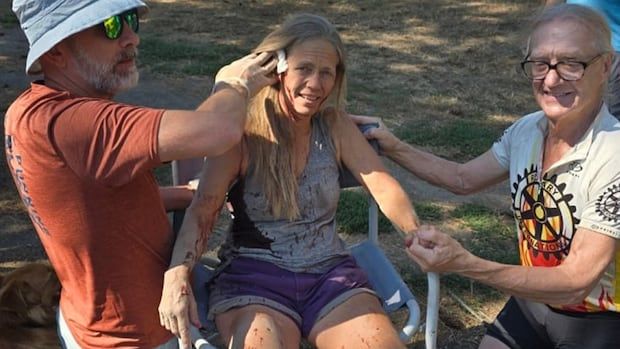 A woman with blood splatter on her sits in a chair while a man holds a bandage to her head and another holds her hand. 