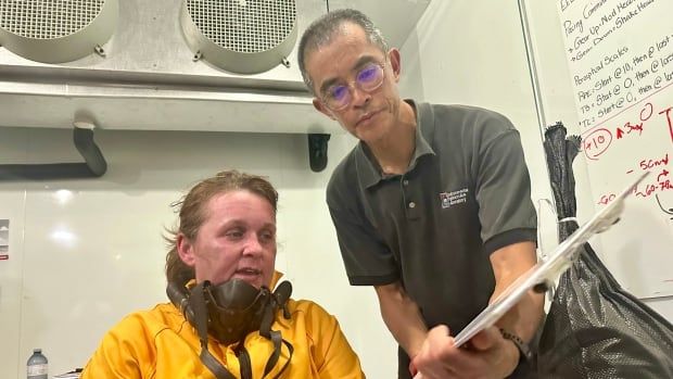 A woman wearing a protective face mask and yellow jacket speaks to an adult man wearing a black tshirt.