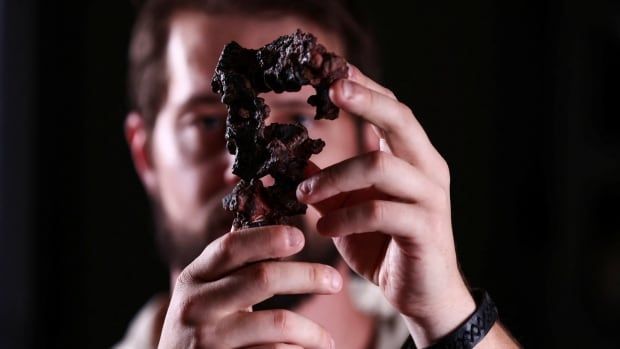 The camera focuses on a twisted lump of dark brown rock and bone, which is being held up by a man directly in front of his blurred, out of focus face. There is a black backdrop to the image. 