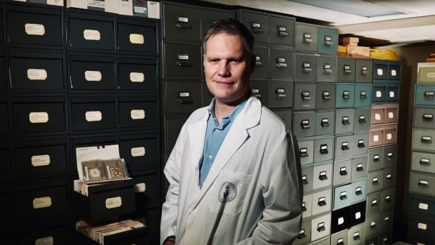 A man in a lab coat stands in front of a wall of black filing cabinets