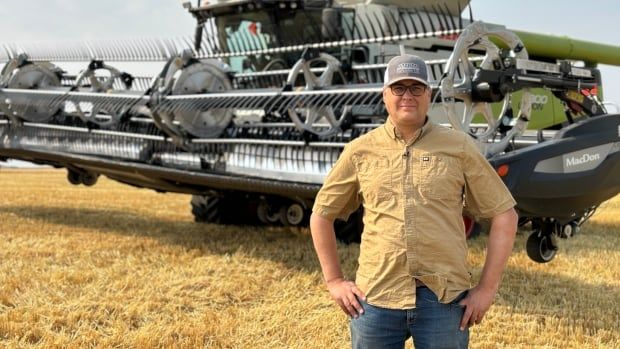 Man in tan shirt, jeans and baseball cap stands in field in front of grain combine with hands on hips