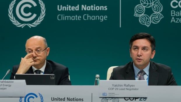 Two men sit at a panel in front of a teal backdrop that says United Nations Climate Change. The man on the left is older and scratching his nose, looking bored. The man on the right appears to be in the middle of talking. 