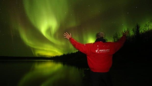 Seen from behind, a man waves his arms joyfully at the sky, filled with swirling green and red aurora borealis.