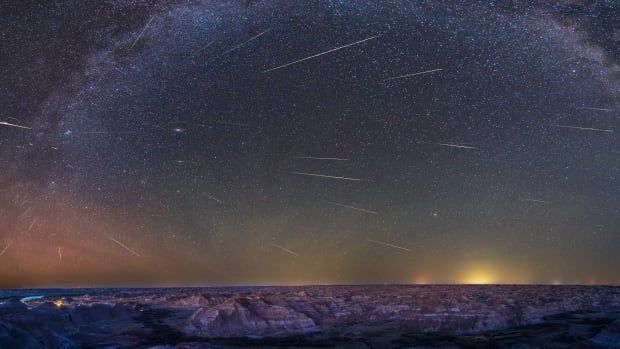 Streaks of light are seen against a star-studded sky, with old rock formations below.