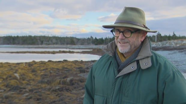 A man in a coat and hat stands on a Nova Scotia shoreline. 