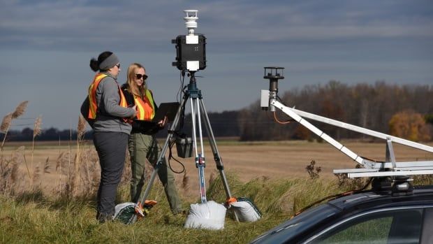 Two women in high-vis safety vests stand behind a tri-pod while they look at a laptop. 