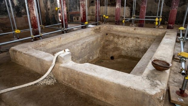 A beige stone rectangular basin is seen in the centre of a room, with red pillars around the outside of it. Metal scaffolding is seen around the bath. 