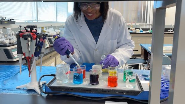 A female researcher wearing a lab coat and goggles looks over beakers of multi-coloured samples.