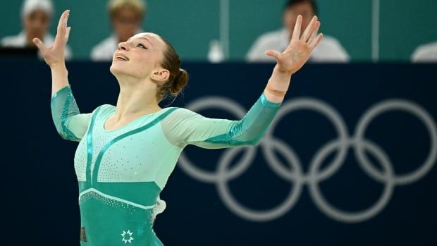 A woman in a leotard holds her hands up and smiles. Olympic rings are visible in the background.