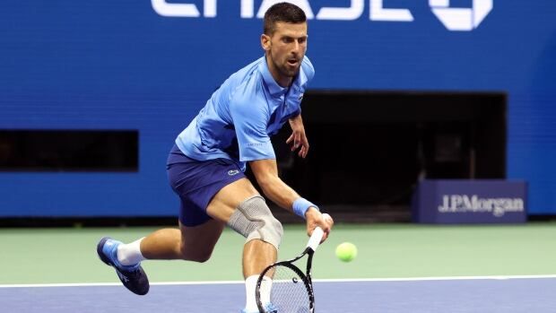 A male tennis player returns a low shot with a racket in his right hand on a hard court.