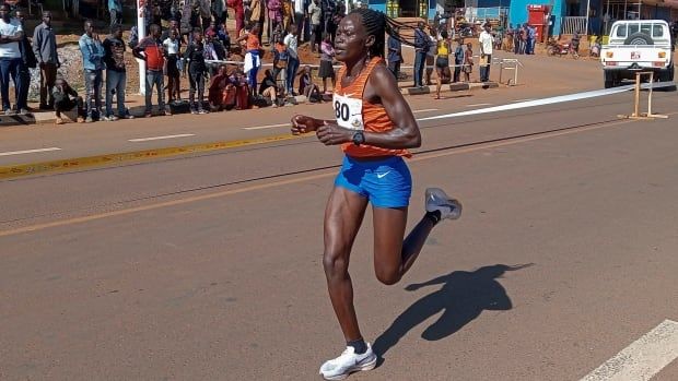 A woman, wearing an orange tank top and blue running shorts, runs on a road past a crowd of people. 