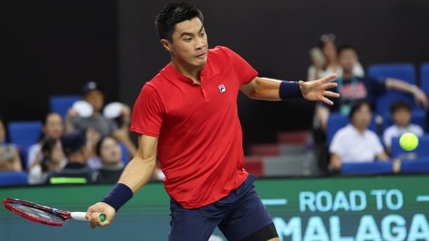 A male tennis player wearing wrist bands swings a racket in his right hand as a ball flies toward him through the air.