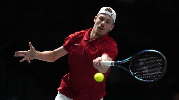 A male tennis player wearing a backwards hat returns a shot with the racket in his left hand.