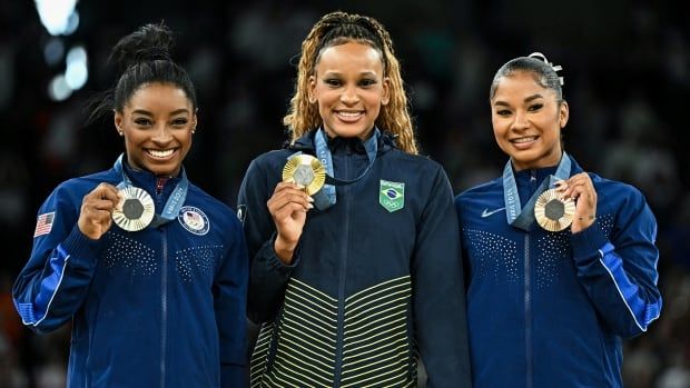 Three female artistic gymnasts wearing track suits smile while holding up medals that hang around their necks.