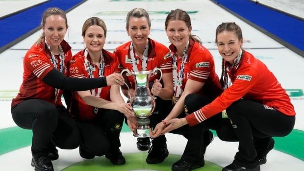 Five female curlers smile while posing with a trophy on the ice.