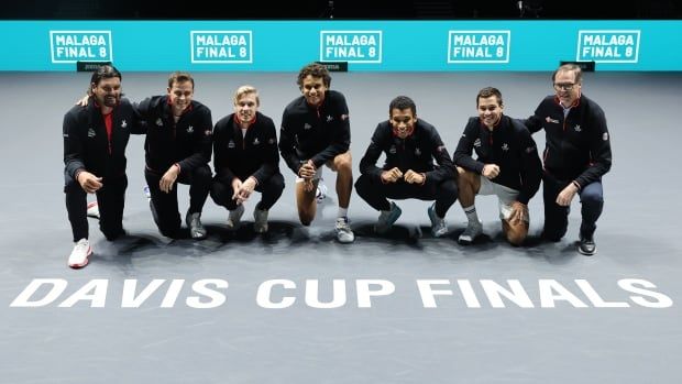 Seven men crouch down on a tennis court in front of a graphic that says 'DAVIS CUP FINALS' in white lettering.