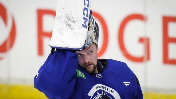 A goalie in blue training gear with the Canucks logo lifts his mask with a padded hand with a blurred-out Rogers logo on the white boards behind him.