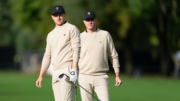 Two male golfers stand side by side as they look off into the distance on a course during the day.