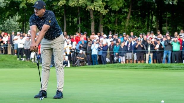 A male golfer leans on his putter while looking dejected as spectators look on from behind a barricade to his rear.