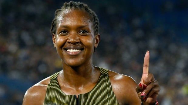 A female track athlete smiles while holding up her left index finger.